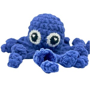 May include: A handmade, plush blue octopus toy with white and black eyes. The octopus has eight textured tentacles and a rounded head. The toy is made of soft, fluffy yarn, suitable for children or as a decorative item.