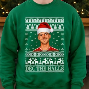 May include: Green Christmas sweater with a festive design. The sweater features a portrait of a person wearing a Santa hat, surrounded by white Christmas trees, snowflakes, and reindeer. The text "DEC THE HALLS" is printed at the bottom.
