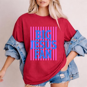 May include: A maroon t-shirt with the text "BIG JESUS FAN" in blue letters, set against a background of pink vertical stripes. The shirt has a relaxed fit.