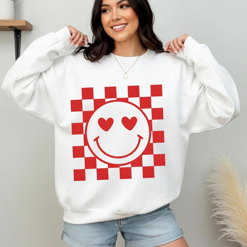 Op de afbeelding: Witte sweatshirt met een rood-wit geblokt patroon en een smiley-ontwerp. De smiley heeft hartvormige ogen en een gebogen mond. De sweatshirt is een casual, comfortabele stijl.