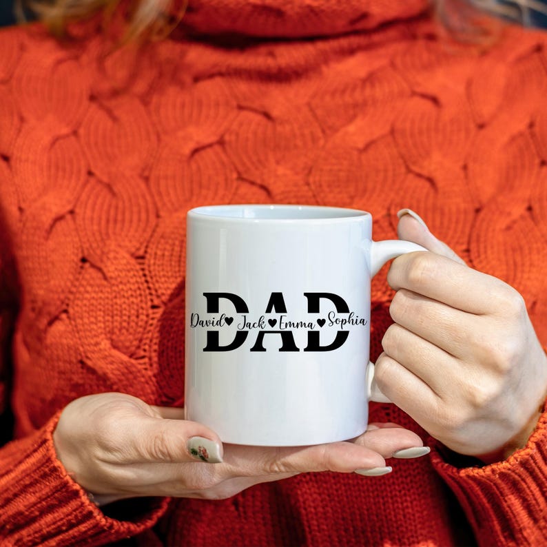 May include: White ceramic mug with the word "DAD" in bold black letters. Below the word "DAD" are the names "David Jack Emma Sophia" in a script font. The names are separated by heart symbols.
