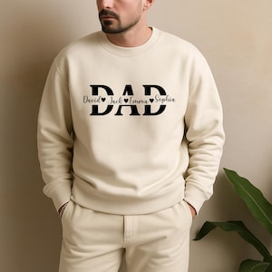 May include: Cream-colored sweatshirt with the word "DAD" in bold black letters. Below the word "DAD" are the names "David", "Jack", "Emma", and "Sophia", each separated by a heart symbol. The sweatshirt is a casual style.