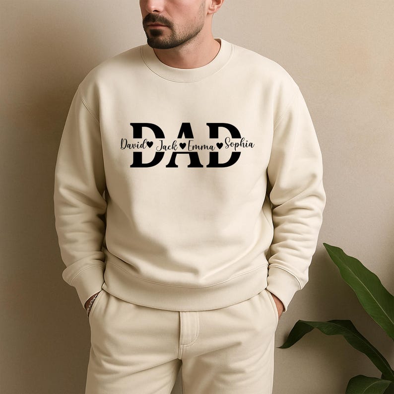 May include: Cream-colored sweatshirt with the word "DAD" in bold black letters. Below the word "DAD" are the names "David", "Jack", "Emma", and "Sophia", each separated by a heart symbol. The sweatshirt is a casual style.