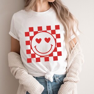 Op de afbeelding: Wit T-shirt met een rood-wit geblokt patroon en een smiley-gezicht ontwerp. De smiley heeft hartvormige ogen en een gebogen mond. Het shirt is gemaakt van een zacht materiaal.
