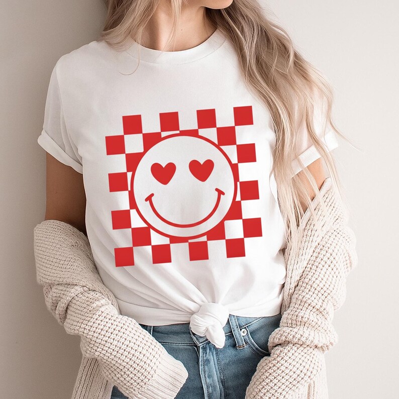Op de afbeelding: Wit T-shirt met een rood-wit geblokt patroon en een smiley-gezicht ontwerp. De smiley heeft hartvormige ogen en een gebogen mond. Het shirt is gemaakt van een zacht materiaal.