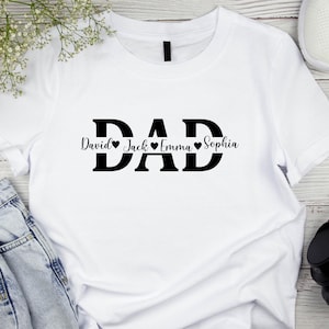 May include: White t-shirt with the word "DAD" in bold black letters. Below the word "DAD" are the names "David", "Jack", "Emma", and "Sophia", each with a heart symbol. The shirt is made of a soft material.