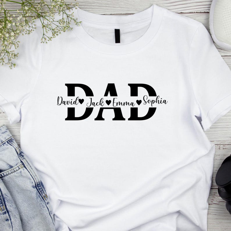 May include: White t-shirt with the word "DAD" in bold black letters. Below the word "DAD" are the names "David", "Jack", "Emma", and "Sophia", each with a heart symbol. The shirt is made of a soft material.