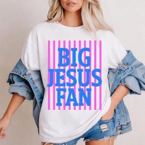 May include: White t-shirt featuring the blue text "BIG JESUS FAN" over pink vertical stripes. The casual outfit includes a denim jacket and shorts. The design is a bold statement.