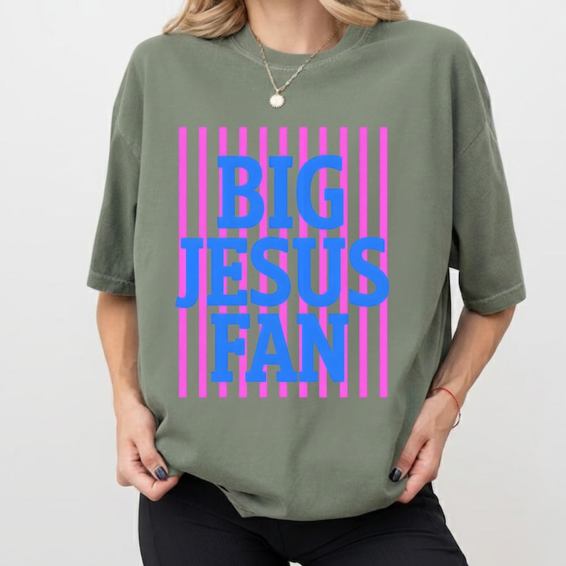 May include: An olive green t-shirt featuring the words "BIG JESUS FAN" in blue block letters, framed by vertical pink stripes. The shirt has short sleeves and a loose fit.