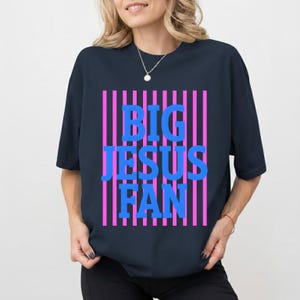 May include: A navy blue t-shirt featuring the text "BIG JESUS FAN" in blue block letters set against pink vertical stripes. The shirt has a crew neck and short sleeves.
