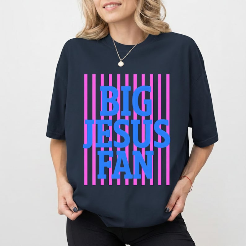 May include: A navy blue t-shirt featuring the text "BIG JESUS FAN" in blue block letters set against pink vertical stripes. The shirt has a crew neck and short sleeves.