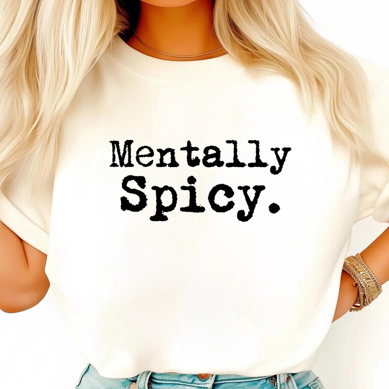 May include: Cream-colored t-shirt with the phrase "Mentally Spicy" printed in black, bold, typewriter-style font. The shirt has a crew neck and short sleeves. The model is wearing blue jeans and gold bracelets.
