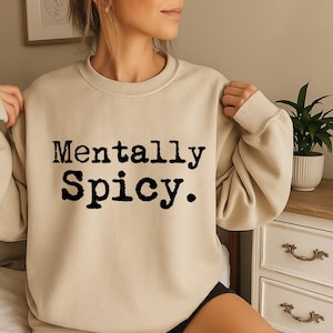 May include: Beige crewneck sweatshirt with the text "Mentally Spicy" in a bold, black font. The sweatshirt is a neutral color, and the text is centered on the front. The style is casual and comfortable.