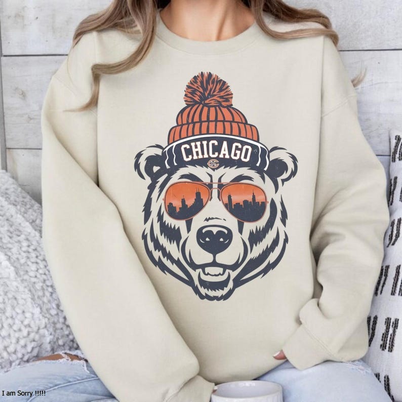 Może przedstawiać: Beżowa bluza z nadrukiem niedźwiedzia w okularach przeciwsłonecznych i czapce z napisem "CHICAGO". Okulary przeciwsłoneczne niedźwiedzia odbijają panoramę miasta. Czapka ma pompon. Bluza jest w neutralnym kolorze.