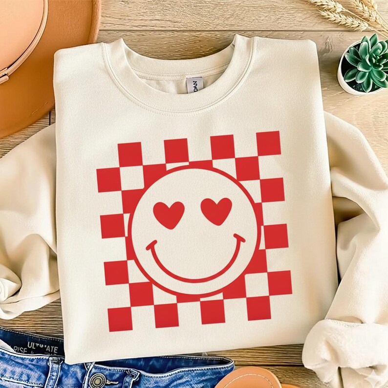 Op de afbeelding: Cr&egrave;mekleurige sweatshirt met een rood dambordpatroon en een smiley-gezichtontwerp. De smiley heeft hartvormige ogen en een gebogen glimlach. De sweatshirt is opgevouwen, met de mouwen zichtbaar.