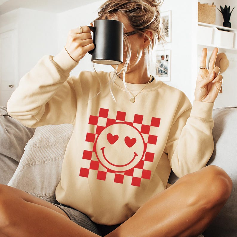 Op de afbeelding: Een cr&egrave;mekleurige sweatshirt met een rood-wit geblokt patroon en een smiley-gezicht ontwerp. De smiley heeft hartvormige ogen. De sweatshirt heeft lange mouwen en een ronde hals.