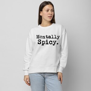 May include: White sweatshirt with the text "Mentally Spicy" in black, bold font. The sweatshirt is a crew neck style and appears to be made of a soft material. The model is wearing light blue jeans.