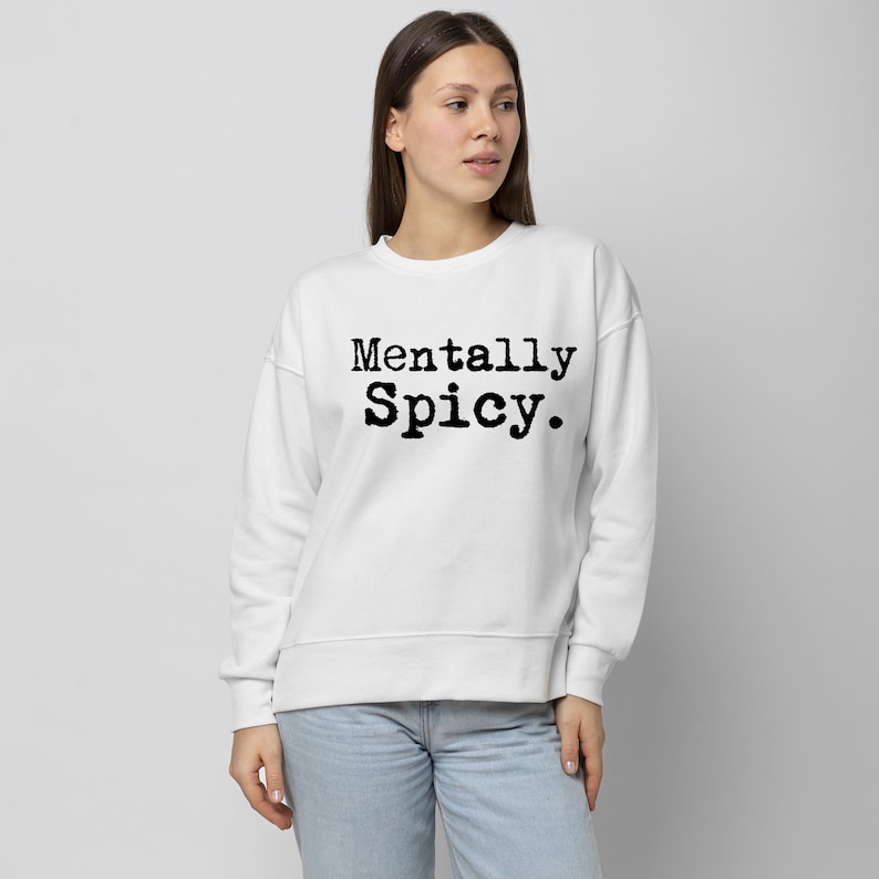 May include: White sweatshirt with the text "Mentally Spicy" in black, bold font. The sweatshirt is a crew neck style and appears to be made of a soft material. The model is wearing light blue jeans.