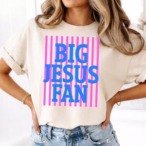 May include: A cream-coloured t-shirt featuring the text "BIG JESUS FAN" in blue, set against a pink striped background. The shirt has short sleeves and a relaxed fit.