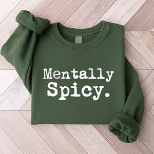 May include: A dark green sweatshirt with the phrase "Mentally Spicy" printed in white, bold, uppercase letters. The sleeves are rolled up, and the sweatshirt is folded neatly. The background is a light wood grain.