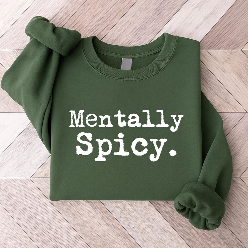 May include: A dark green sweatshirt with the phrase "Mentally Spicy" printed in white, bold, uppercase letters. The sleeves are rolled up, and the sweatshirt is folded neatly. The background is a light wood grain.