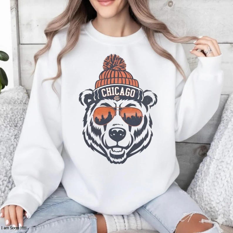 Może przedstawiać: Biała bluza z grafiką niedźwiedzia w pomarańczowej czapce i okularach przeciwsłonecznych ze skyline'em Chicago w soczewkach. Słowo "CHICAGO" znajduje się nad głową niedźwiedzia. Niedźwiedź ma czarno-białe futro.