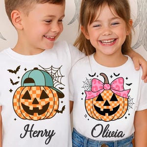 Könnte beinhalten: Zwei Kinder tragen weiße T-Shirts mit Halloween-Kürbis-Designs. Ein Kürbis hat eine blaue Kappe, der andere eine rosa Schleife. Beide Kürbisse haben ein kariertes Muster, ein geschnitztes Gesicht und sind von Fledermäusen und Spinnweben umgeben. Auf dem Shirt des Jungen steht "Henry", auf dem des Mädchens "Olivia".