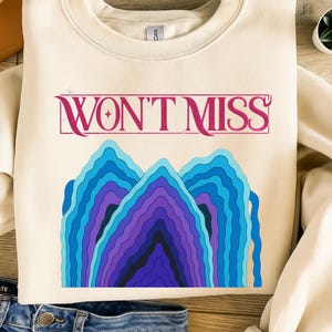 May include: Cream-colored sweatshirt featuring the text "WON'T MISS" in a pink, stylized font above a graphic of layered blue and purple mountain shapes. The sweatshirt is paired with blue denim jeans.