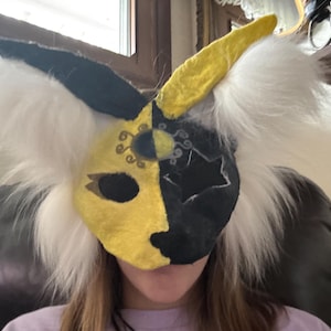 May include: A handmade, half-face mask with a split design: yellow and black. The mask has white, fluffy fur around the edges and a yellow horn. The mask is designed with decorative cutouts and is likely for cosplay or costume use.