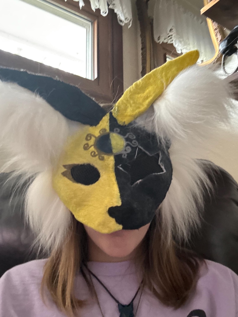 May include: A handmade, half-face mask with a split design: yellow and black. The mask has white, fluffy fur around the edges and a yellow horn. The mask is designed with decorative cutouts and is likely for cosplay or costume use.