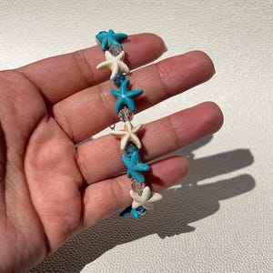 Könnte beinhalten: Ein Perlenarmband mit abwechselnd weißen und türkisfarbenen Seestern-Anhängern. Klare Perlen sind zwischen den Seesternen eingefügt. Das Armband wird in einer Hand gehalten und präsentiert den Schmuck mit Strandmotiven.