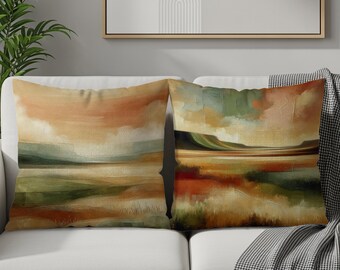 Puede incluir: Dos cojines decorativos con arte abstracto de paisaje. Los cojines presentan una paleta de colores naranja, verde y marrón, que evoca una puesta de sol sobre un campo. Los cojines son rectangulares y descansan sobre un sofá blanco.