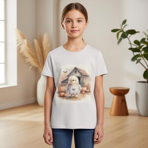 May include: White t-shirt with a Halloween graphic. The design shows a chick in a ghost costume in front of a barn with pumpkins. The t-shirt is worn by a person.