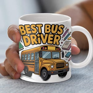 Puede incluir: Taza de cerámica blanca con una ilustración de autobús escolar amarillo y el texto "BEST BUS DRIVER". El diseño incluye libros y lápices. La taza está sujeta por una persona.