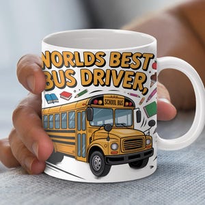 Puede incluir: Taza de cerámica blanca con el texto "Worlds Best Bus Driver" en amarillo y negro. Un gráfico de autobús escolar amarillo aparece en la taza, junto con ilustraciones de libros y lápices. La taza está sostenida por una persona.