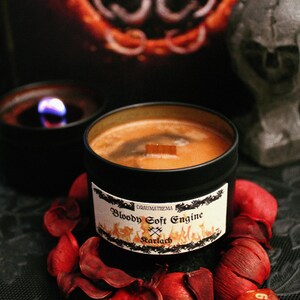 Può includere: Un barattolo di candela nero con uno stoppino di legno e un'etichetta che recita "Draumathema Bloody Soft Engine Rarlach". La candela è circondata da petali di fiori rossi e un dado rosso e arancione.