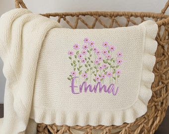 May include: Cream-coloured baby blanket with a floral embroidery design and the name "Emma" in purple script. The blanket has a ruffled edge and is displayed in a woven basket.