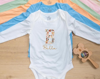 May include: A collection of long-sleeved baby onesies in various pastel colors, including white, blue, pink, and peach. The white onesie features a floral letter "B" and the name "Bella". A wooden baby rattle is also visible.