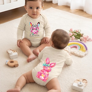 May include: Two babies wearing cream-colored onesies with Easter bunny designs. One features a bunny face with glasses and the name "Olivia." The other has a pink bunny on the back. Toys and a rainbow are nearby.