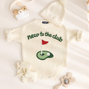 May include: A cream-colored knit long-sleeve romper featuring the embroidered text "new to the club" above a golf green and flag graphic. The set includes a matching cream knit beanie with a pom-pom, a pair of cream knit booties, a wooden toy camera, and a wooden koala-shaped rattle.