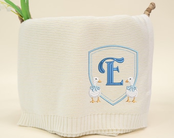 May include: Cream-colored knitted baby blanket with a blue embroidered crest featuring the letter "E" and two ducks. The blanket has a ribbed border and is decorated with a sprig of lily of the valley.