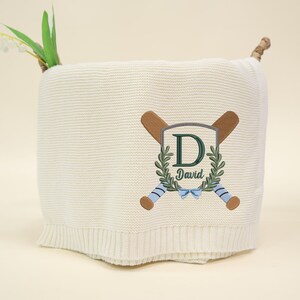 May include: Cream-colored knitted baby blanket with a baseball-themed embroidered design. Features crossed baseball bats, a shield with the letter "D" and the name "David", and a blue bow. The blanket is folded and has a decorative stem.