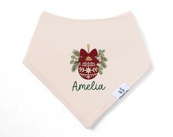 May include: A cream-colored triangular bandana bib featuring a central graphic of a red Christmas ornament with a bow, surrounded by green pine branches and white berries, with the name Amelia printed in dark green script below.