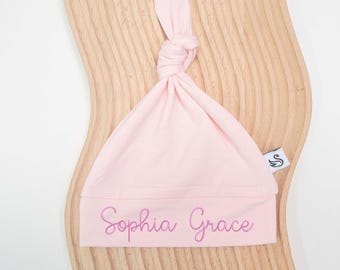 Könnte beinhalten: Hellrosa Baby-Knotenmütze mit dem gestickten Namen "Sophia Grace" in Schreibschrift. Die Mütze ist aus weichem Stoff gefertigt und hat eine geknotete Oberseite. Ein kleines Etikett mit einem Schwan-Logo ist angebracht.