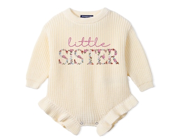 May include: A cream-colored long-sleeve knit romper with a ribbed texture, featuring a ruffled hem and a crew neckline. The chest displays the text 'little' in pink script above the word 'SISTER' in a floral pattern with pink and green tones.