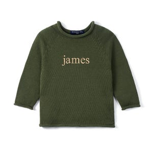 May include: Olive green knit sweater with the name "james" embroidered in a light tan thread across the chest. The sweater has a crew neck and long sleeves. The brand name "Swaddle AN..." is visible on the back of the neck.