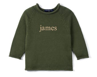 May include: Olive green knit sweater with the name "james" embroidered in a light tan thread across the chest. The sweater has a crew neck and long sleeves. The brand name "Swaddle AN..." is visible on the back of the neck.