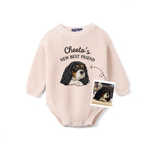 May include: A light pink long-sleeve knit baby bodysuit featuring an embroidered Cavalier King Charles Spaniel and the text Cheeto's NEW BEST FRIEND. A small rectangular photograph of the same dog is attached to the front of the garment.