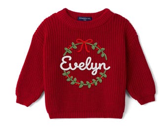 May include: A deep red long-sleeve knit sweater featuring a crew neckline and a ribbed texture. The center displays an embroidered green wreath with red berries, a red bow, and the white script text Evelyn. The inner label reads Swaddle AN.
