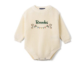 May include: A cream-colored long-sleeve knit romper featuring a ribbed texture and a crew neckline. The chest displays the embroidered name Brooks in green, flanked by two flying ducks holding a string of colorful holiday lights.
